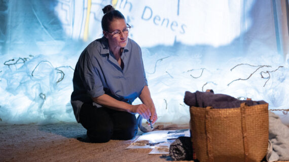A woman kneels on the floor. An image is projected brightly behind her. She is looking at a basket
