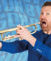 Jason Byrne Irish Male comedian holding a trumpet. He has his mouth open as he looks directly at the camera. He is wearing a blue suit. The background is blue. He is holding a fake head at the end of the trumpet. The event is called Jason Byrne Unblocked.