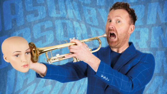Jason Byrne Irish Male comedian holding a trumpet. He has his mouth open as he looks directly at the camera. He is wearing a blue suit. The background is blue. He is holding a fake head at the end of the trumpet. The event is called Jason Byrne Unblocked.