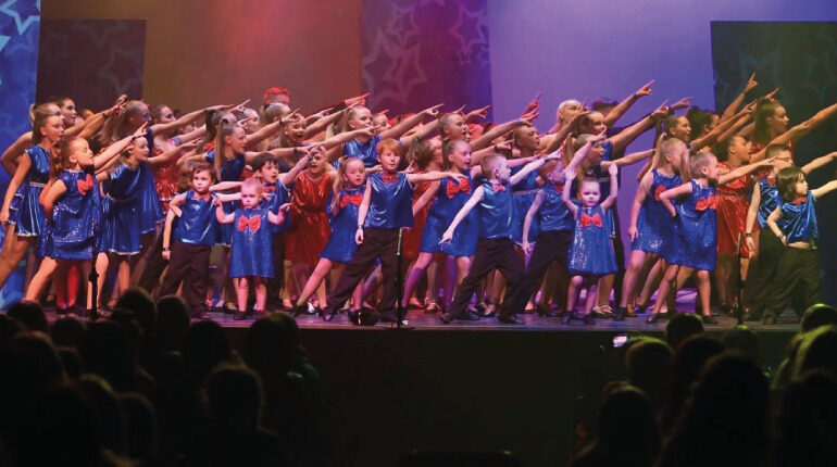 Children performing on lit stage in a variety show. The children are dancing and wearing blue outfits. We can see the back of the audiences head. The dance group is part of Dance City Ireland.