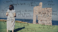 A Woman beside the sea with her back to the camera. It is a sunny day. She is looking at a castle. The screen has writing etched over it.